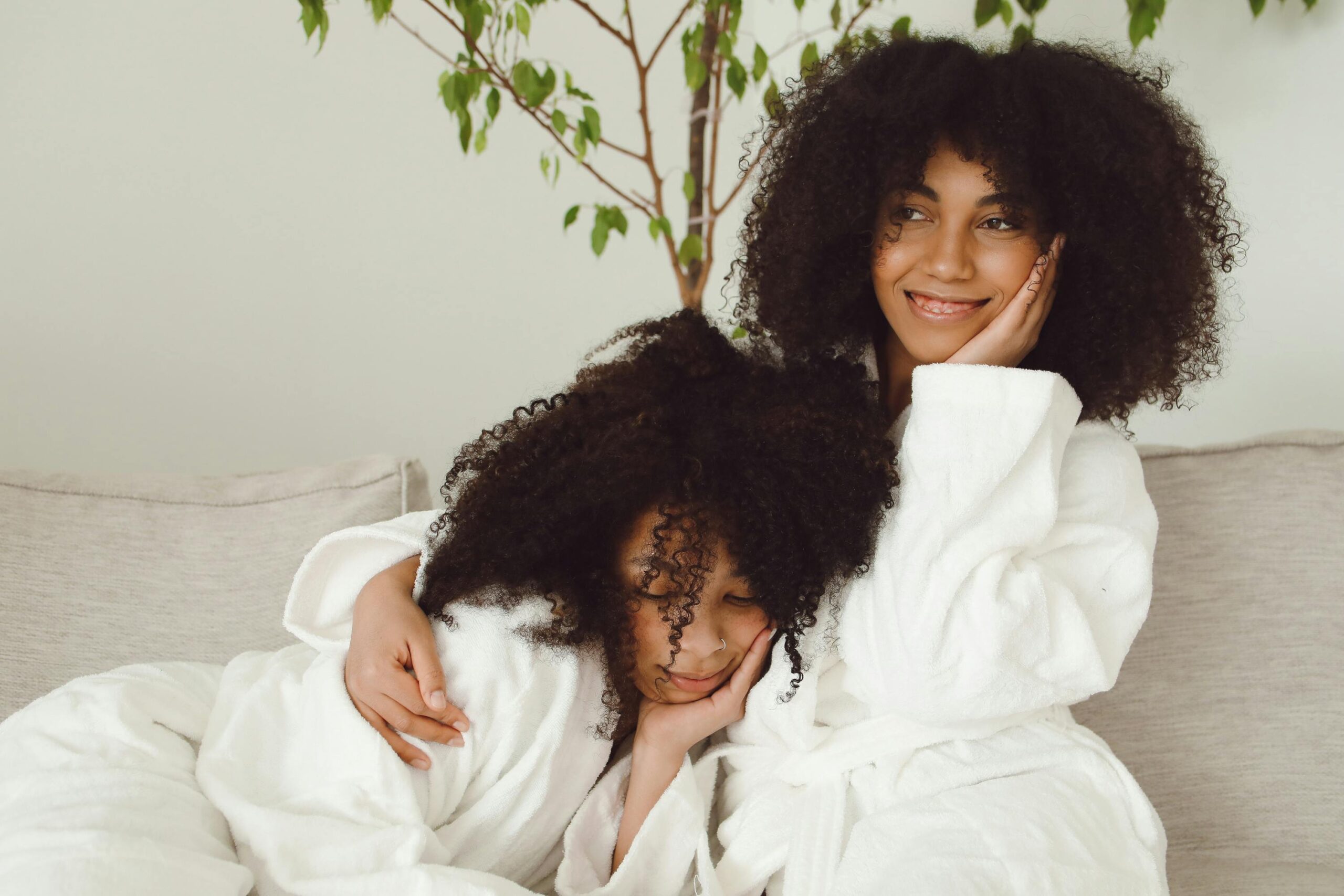 Two smiling women in bathrobes enjoying a relaxed moment on a sofa with lush green plant nearby.