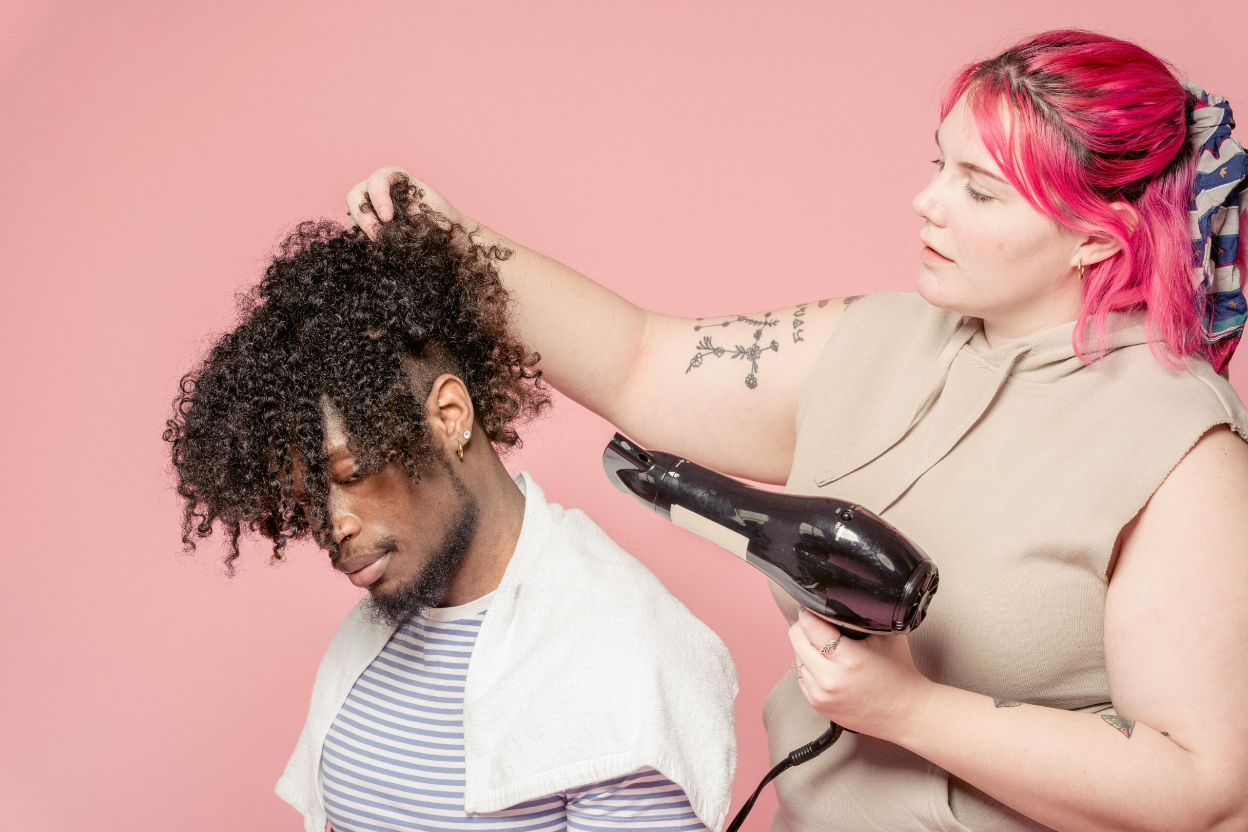 Professional hairdresser with dyed bright hair drying hair of African American male customer while working against pink background