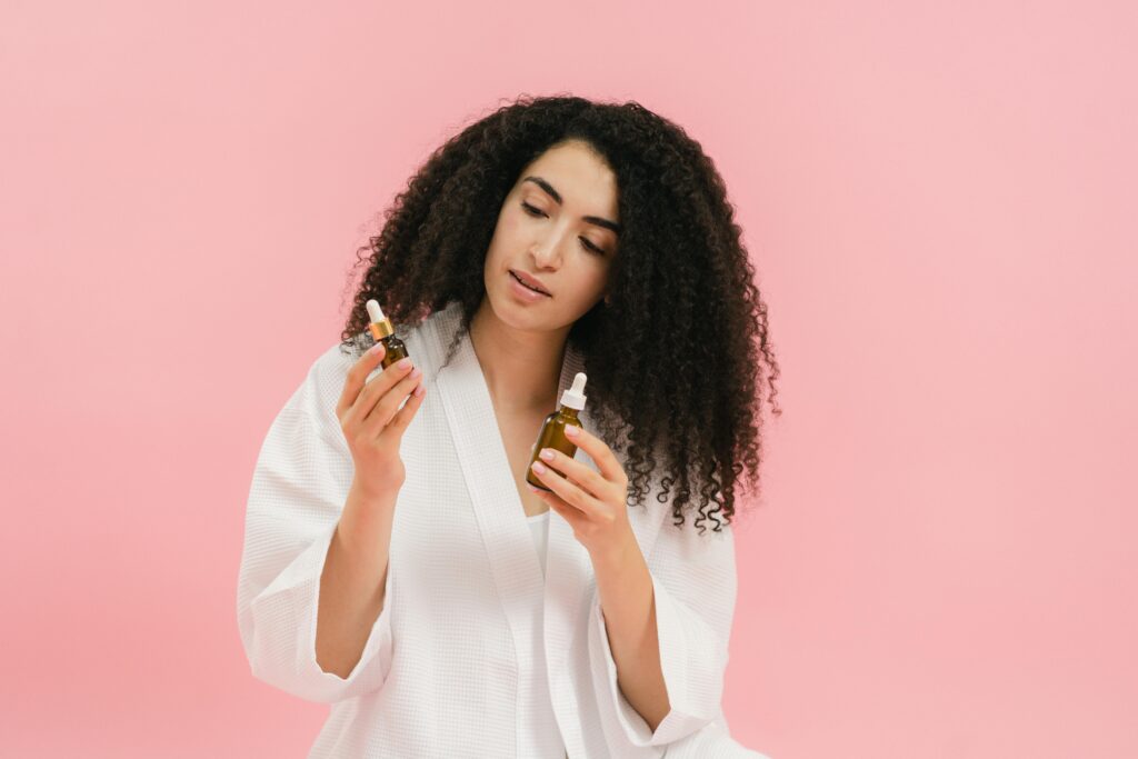 A woman with curly hair in a white robe examines two cosmetic oil bottles against a pink background.